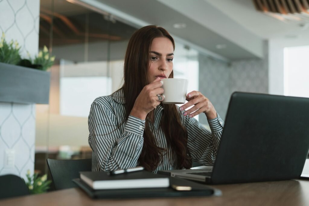 Young woman drinking coffee while working on laptop in a modern office setting, focusing on productivity.