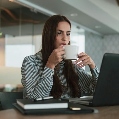 Young woman drinking coffee while working on laptop in a modern office setting, focusing on productivity.